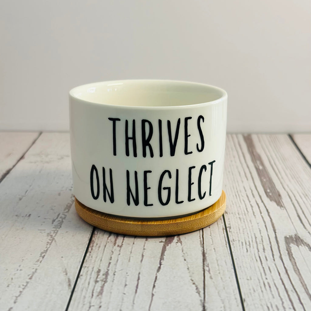 White pot with black text 'THRIVES ON NEGLECT' on a wooden surface