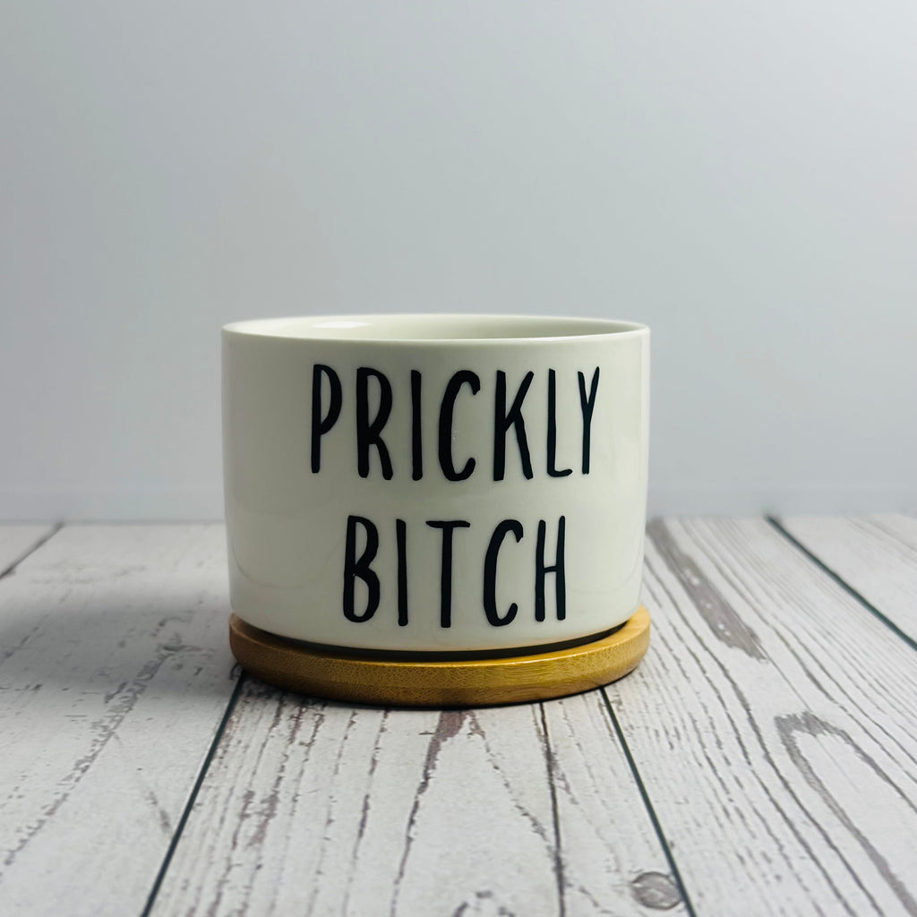 White pot with 'PRICKLY BITCH' text on a wooden surface