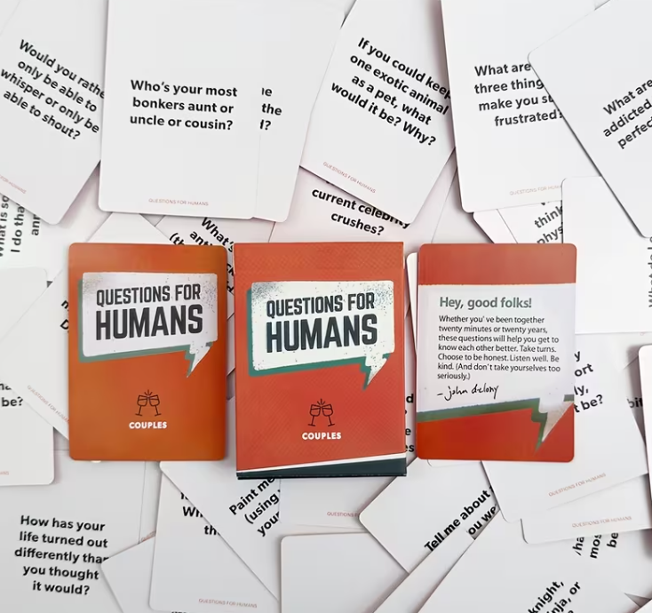 A set of 'Questions for Humans' couples cards scattered on a white background, showing orange and white card faces with printed text prompts.